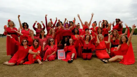 John Jones A large group of women wearing red dresses, some with their arms up, all are smiling and looking at the camera. They are sitting or standing on grass.