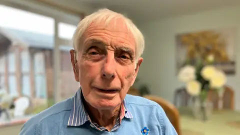 Brian Standring sits in a room at the Ivy House Sheltered Housing Home - he wears a striped shirt and a blue jumper with a flower pin