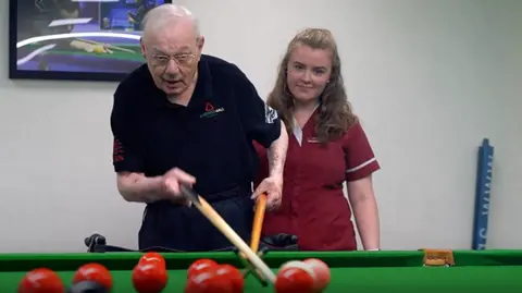 An elderly man in a black polo short take a snooker shot using a rest whilst a young woman in a maroon uniform looks on.