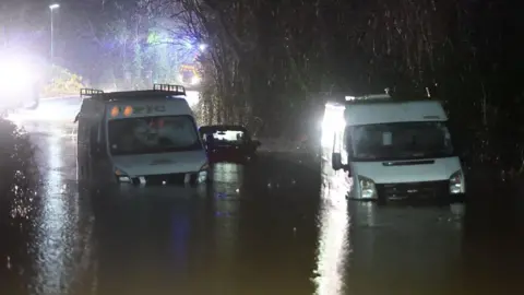 Two white vans and a dark coloured car in between them sit in deep water almost up to the bonnets. Bright lights from emergency vehicles shine in the background in the darkness