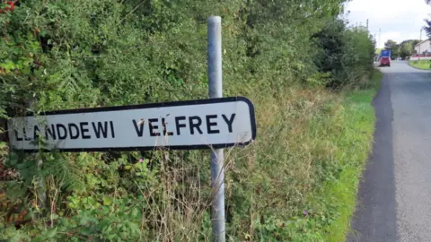 A sign of Llanddwei Verfrey. It's a white sign with black writing in a green bush next to a road. 