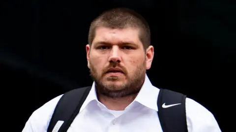 Zak Archbold pictured leaving court. He has short brown hair and a beard. He is wearing a white shirt and has a black backpack over his shoulders.