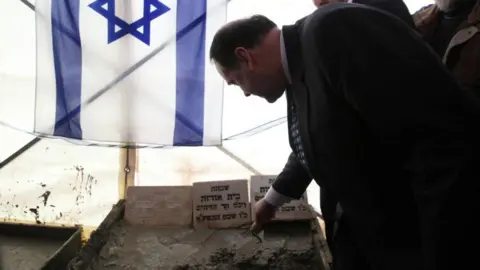 AFP Former Arkansas politician  Mike Huckabee adds factual  to a country   chromatic  successful  beforehand   of an Israeli emblem  during a dedication ceremonial  for a caller   Israeli colony  astatine  Beit Orot, successful  occupied East Jerusalem connected  31 January 2011.