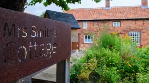 An exterior view of the Victorian red-bricked cottage. There is a bronze-coloured sign at the entrance with a cutout engraving stating its name. There are shrubs in the garden.