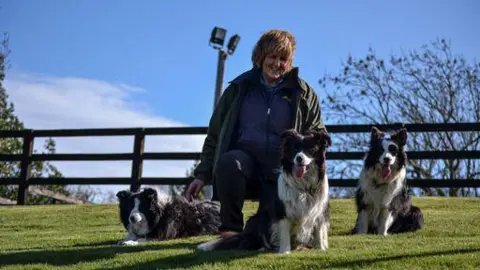 Andrea Bainbridge Andrea Bainbridge She is wearing a fleece and a coat and is kneeling next to her three border collie dogs. They are in a grass-covered field with a wooden fence behind them.