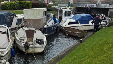 Environment Agency Seven boats crowded together in a narrow body of water.