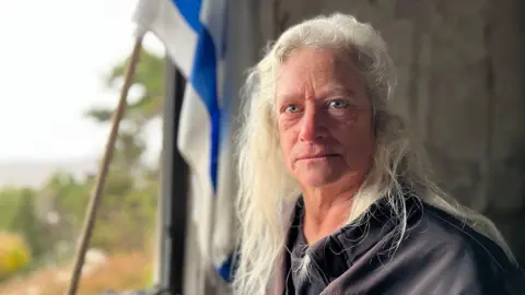 Kibbutz Menara resident Orna. She is seen standing in front of an empty window frame with pieces of debris on the windowsill. An Israeli flag is propped up inside the frame. Orna has long grey hair and is looking straight into the camera. 