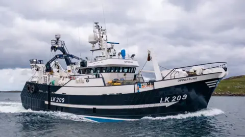 The fishing boat is white and blue and has the ID "LK209" and its name "Opportune" on its hull. The boat is leaving a frothy wake as it travels across the sea near an area of low-lying shoreline.