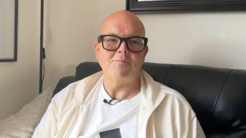 BBC Jody Bunting sitting on the sofa wearing glasses, a white T-shirt and cream zip-up top