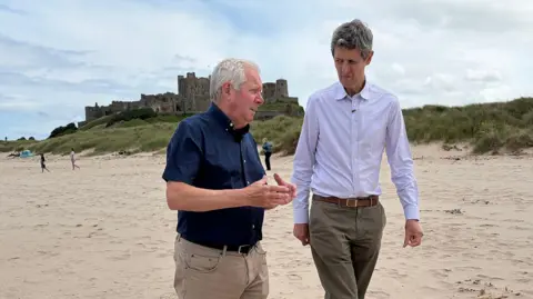 Two men in their 60's are walking down a wide sandy beach. In the background is Bamburgh castle and there are a few clouds in the sky. The sea is calm and the men are clearly engaged in conversation. Both are wearing trousers and relaxed shirts.