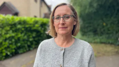 Stephen Huntley/BBC A woman wearing a grey cardigan with buttons and a silver necklace. She has blonde cropped hair down to her neck, and grey-rimmed glasses. She is standing in front of a house with a hedge running in front of it.