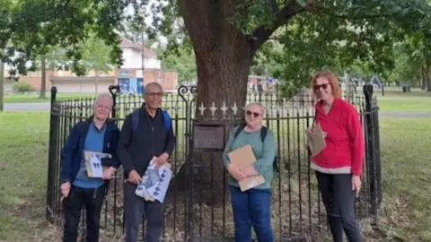 Four tree volunteers stood in front of a well-established oak during the summer months, which is fenced off and has a plaque