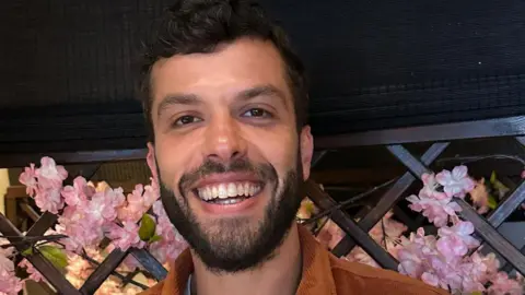Aziz Ziriat , a young man with a dark beard and hair smiles as he looks into the camera. There are blossoms in the background of the image. 