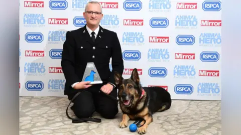 PA Media PD Finn, winner of the Hero Animal of the Year award in 2017, with his handler - former PC Dave Wardell - crouched down and posing for the camera at the Grosvenor House Hotel, London, in front of a billboard with sponsors on it.