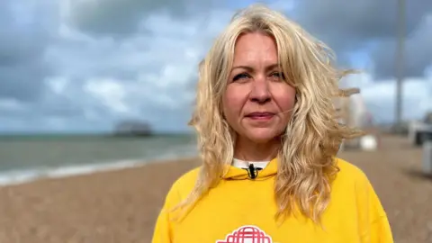 A woman with long blonde hair wearing a yellow hoodie with a red image of Brighton's West Pier. She is stood looking into the camera while stood on the pebbles of Brighton beach