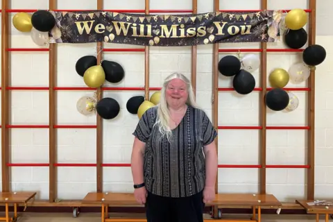 Judith Harrison standing in front of a black and gold banner that says 'we will miss you surrounded by gold and black balloons