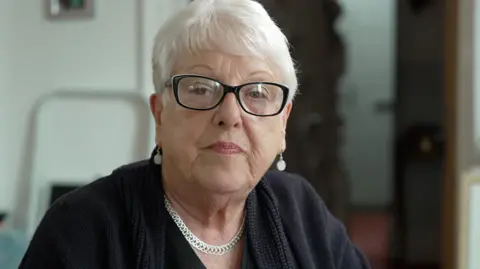 A woman with short white hair, glasses and a dark black top looks serious as she faces the camera. She is also wearing a silver-coloured necklace and earrings.