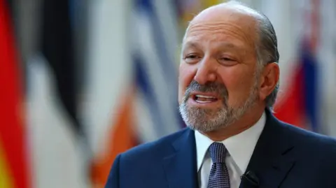 Reuters El secretario de Comercio de Estados Unidos, Howard Lutnick, vestía un traje azul marino. camisa blanca y corbata azul de lunares De pie frente a una bandera nacional que no está en el centro de atención
