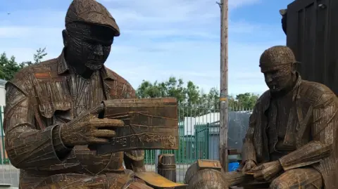 Handout Two sculptures of men - one is wearing a flat cap and reading a newspaper. The other is wearing a helmet.