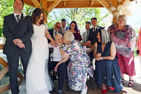 Richard Swingler Wedding celebrations including a bride and groom on the left and other family members wearing dresses and suits. The group are in some kind of wooden gazebo with trees seen in the background. 
