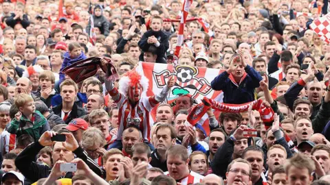 Getty Images crowds of fans on the pitch after beating Coventry, they are singing, clapping, waving scarves and banners and holding children on their shoulders.