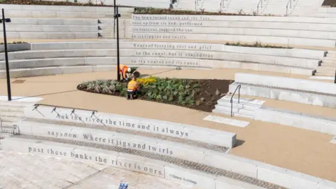 The white stone steps lead to the amphitheatre. There are flower beds dotted around the platforms that break up the steps. The words engraved read: "The town turned round and the Tees; rose up from its bed, performed; its festival, juggled the sun; in its ripples and swirls.; Years of ferrying rust and grief; to the coast, but it stayed true; to its course and its cause; Ask the river the time and it always; says now. Where it jigs and twirls; past these banks the river is ours."