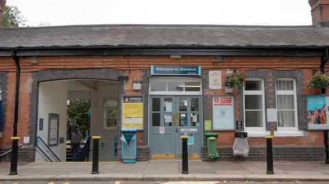 Chiltern Railways The exterior of Warwick railway station. It is a low, red brick building with a double door entrance in the middle of the shot. to the left of the main doors is a set of stairs leading to the subway connecting the north and southbound platforms.