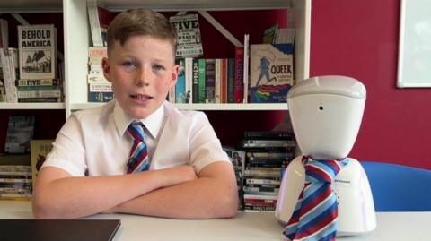 Howard sitting at a desk in school uniform next to AV Howard, a small white tabletop robot wearing a red and blue striped tie