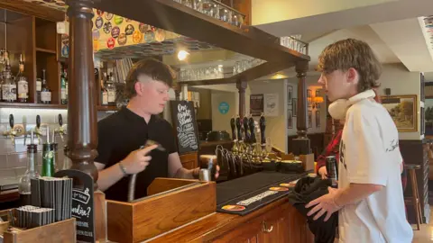 Joshua is filling a pint of coke for a customer who is wearing a white t-shirt with headphones around his neck. 