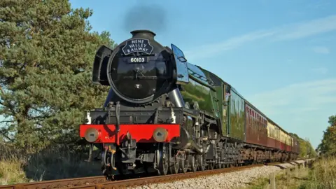 The Flying Scotsman, a green locomotive, pulls carriages along a track through the countryside. Smoke is coming out of the loco's funnel.