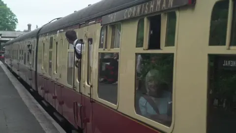 A cream/maroon heritage passenger train stands at a station platform with the name “THE CORNISHMAN” displayed along the side. Several passengers lean out of the carriage windows looking toward the platform, while others stand nearby. Trees and station structures are visible along the platform in the background.