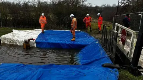 Four men in orange coats and white helmets standing on blue and white-covered ground, looking at an area of water