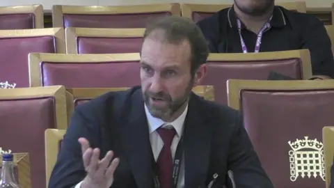 House of Lords Matt Collins, a bearded, middle aged man in a dark suit and red tie, makes a point at a select committee hearing