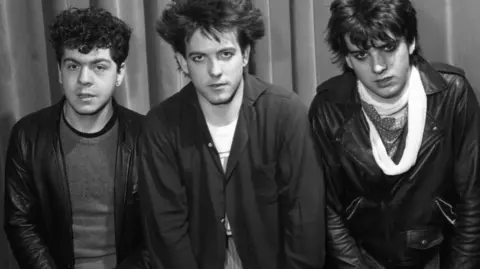 BBC Rock band The Cure, pictured backstage at BBC Top of the Pops in 1981. The image featuring three band members is in black and white, with singer Robert Smith pictured in the centre.  