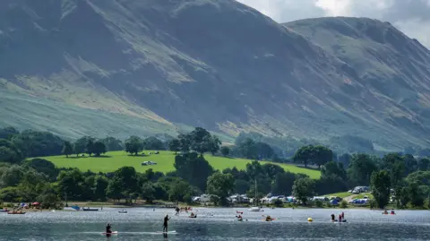 PA Media Crowds enjoying clear weather at Ullswater in the Lake District. The lake is in the foreground with several people paddle boarding, kayaking and swimming. On the other side of the lake there are several cars parked and people on the shore. 
