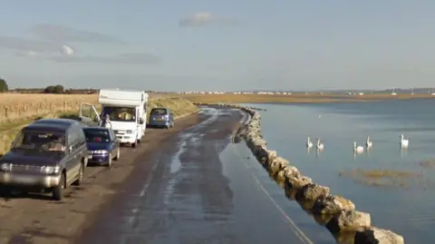 A road alongside a marshy estuary where there are five swans swimming. Parked in a layby on the left is a camper van and three cars.