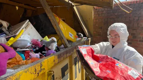 Mark Ansell/BBC A young woman in a white hazmat suit empties a red bag of waste into a full yellow skip