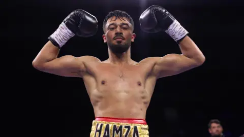 Hamza Uddin wearing boxing gloves and gold lame shorts bearing the word HAMZA. He is standing with his gloves raised in a triumphant pose. 