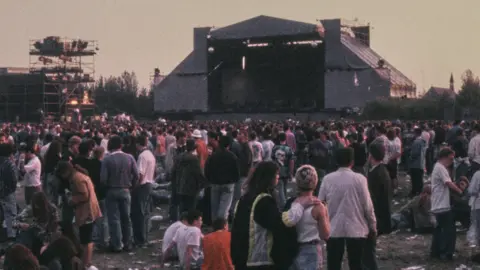 Stone Roses gig on Spike Island in Widnes in 1990. There are countless music fans standing in front of a large pyramid-shaped stage.