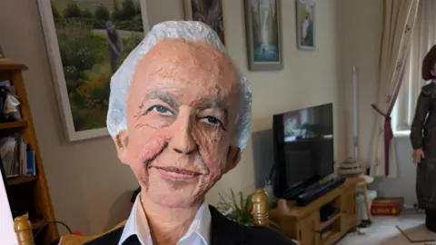 Sharon Kulesa A head and shoulders image of a sculpture depicting Sir David Attenborough wearing a black suit and a white shirt. He is inside a living room. 