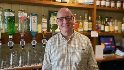 Tom is smiling at the camera. He is wearing black framed glass and a checked shirt.