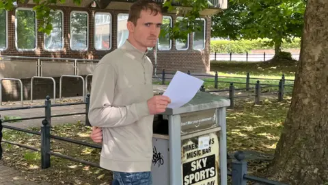 Corey Coleman is walking out of Merhyr Tydfil Magistrates' Court - it is a low, brown brick building and eight, oblong windows can be seen behind him. Coleman is wearing blue denim jeans and a long-sleeve dark cream quarter zip top. Corey has short dark hair and has his left arm behind his back while holding a white piece of paper in his right hand. A bin with a flyer stuck to it is directly next to him and there are trees and grass behind him. Corey has a very stern expression on his face