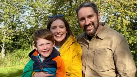 David Metherell Patient David pictured wearing a beige shirt posing for a picture with his wife who is in a yellow jacket and smiling, and his young son who is also smiling in a multi-coloured coat