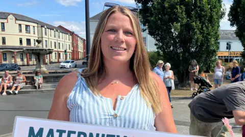 Hannah Rulton holds a sign reading “Maternity Matters” during a public gathering in a town square. Several people stand nearby, with trees, a fountain and buildings visible in the background.