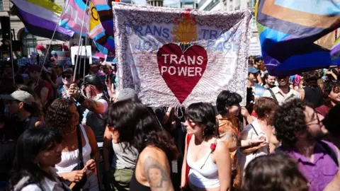Dozens of people are seen gathered in the background and foreground at the march. A banner is held up by some people in the middle of the photo that reads "Trans joy, trans power, trans rage"