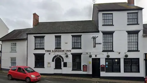 Google Street View image of the London Inn in Ottery St Mary. It is a white brick building with a red car outside on the road. It has several sash windows with black frames. It has a black/grey tiled roof.
