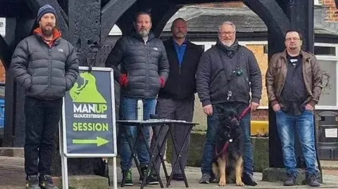 MAN UP Gloucestershire Five men are standing on the pavement. One of them is standing next to a sign that says MAN UP Gloucestershire session, with an arrow pointing to the right below the words. One of the men has a dog on a leash.