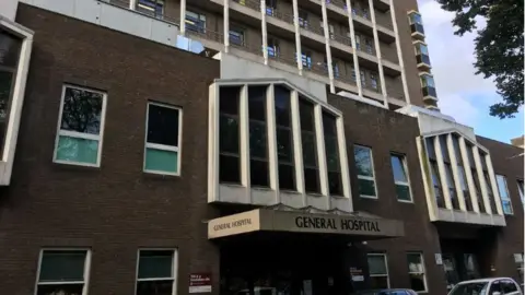 BBC The photo shows the exterior of a multi-storey hospital building. The sign above the entrance clearly reads “GENERAL HOSPITAL.” The architecture features dark brick walls with large, vertical windows framed in white concrete. There are several floors visible, with balconies on the upper levels. At street level, there are cars parked in front of the entrance, and some signage is visible near the doorway. 