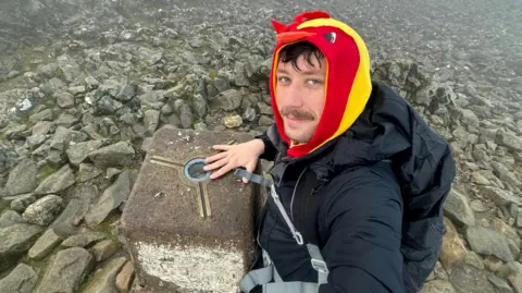 Luke takes a selfie at the summit of one of the mountains. He is wearing a jacket over the top of his chicken costume.
