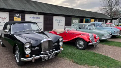 Classic cars in black, red, green and blue are parked up beside a long black building. 